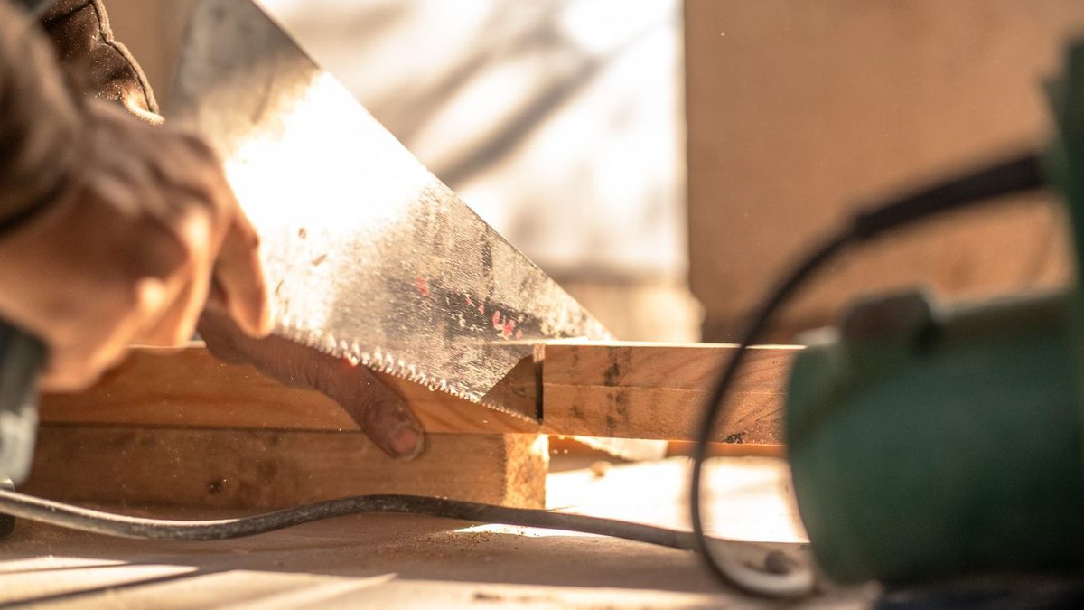 Les mains d'un artisan occupées à scier une pièce de bois, sciure en suspension dans la lumière dorée de l'atelier.
