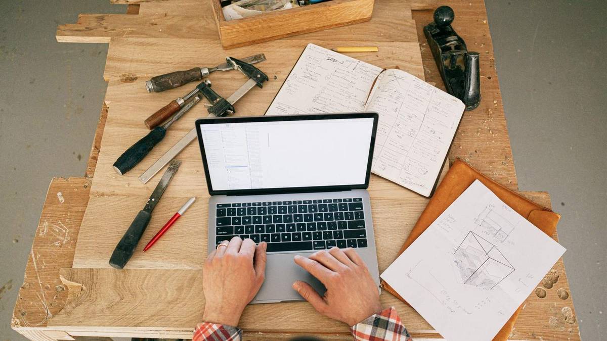 Les mains d'un artisan en chemise à carreaux tapent sur un ordinateur portable posé sur son établi, entre outils de menuiserie, carnet de notes et croquis.