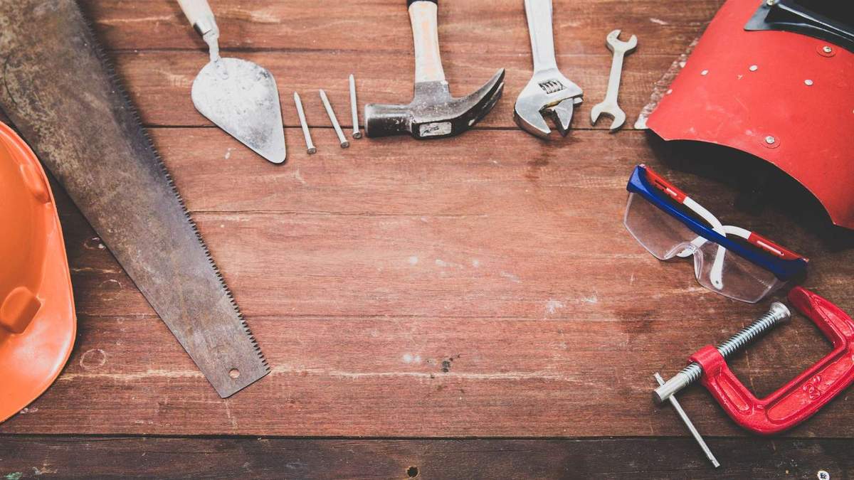 Des outils à main — scie, truelle, marteau, clés, serre-joint — disposés avec soin sur un établi en bois à côté d'un casque de chantier orange.