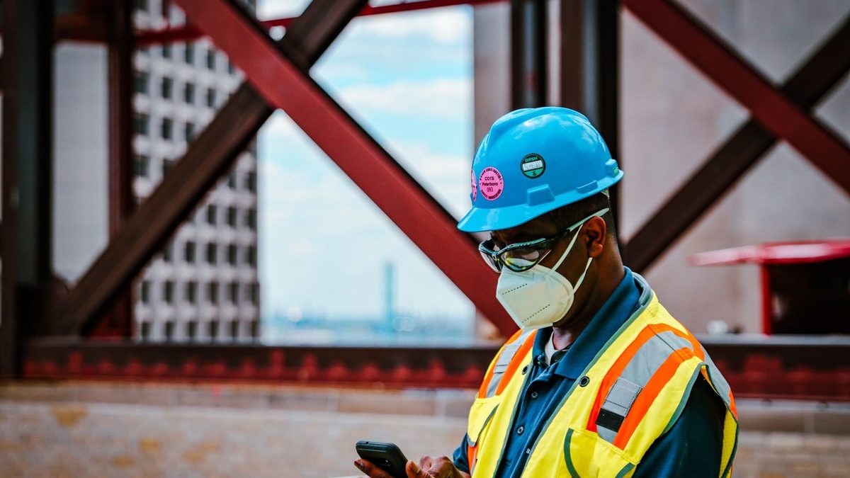 Un ouvrier du bâtiment en gilet jaune et casque bleu consulte son smartphone sur un chantier, devant une charpente métallique.