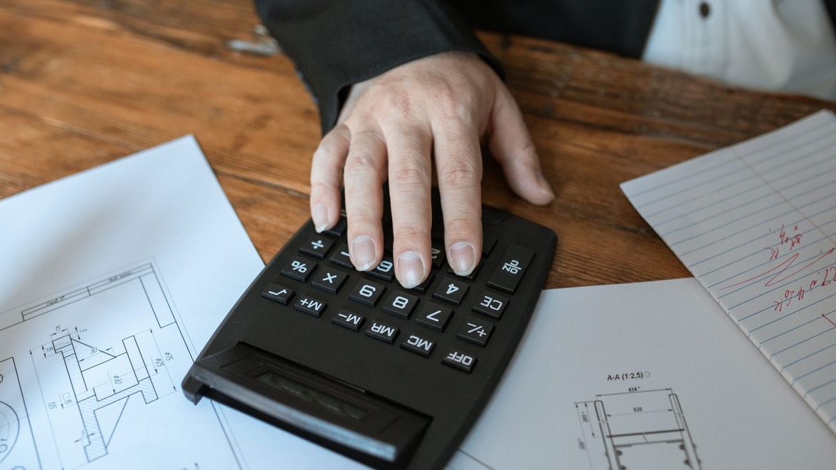 Une main pianote sur une calculatrice posée au milieu de plans techniques et de notes, sur une table en bois.