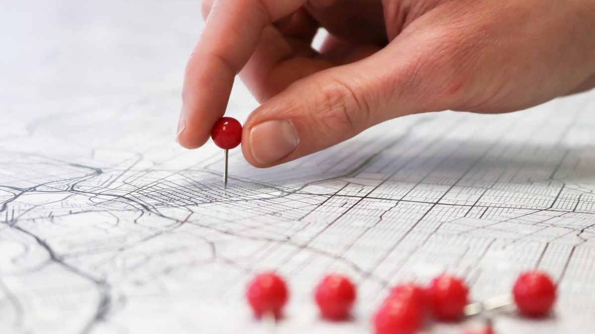 Une main plante une épingle rouge sur un plan de ville détaillé, d'autres épingles posées au premier plan.