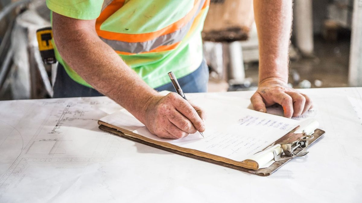 Un ouvrier en gilet haute visibilité prend des notes au stylo sur un porte-bloc posé sur des plans, en plein chantier.
