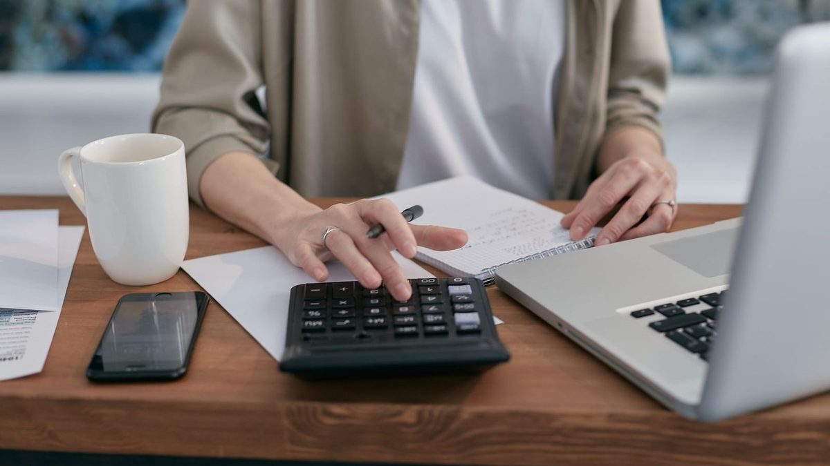 Une personne assise à un bureau en bois compare des chiffres à la calculatrice, un carnet de notes et un ordinateur portable devant elle.